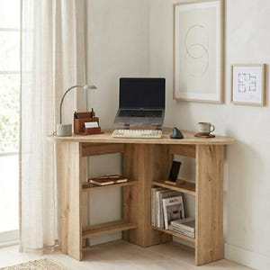 Könnte beinhalten: Ein hellbrauner Eckschreibtisch aus Holz mit eingebauten Regalen. Auf dem Schreibtisch befinden sich ein Laptop, eine Tastatur, eine Maus und eine kleine Lampe. Es gibt einen Schreibtisch-Organizer aus Leder, eine Tasse und gerahmte Kunstwerke.