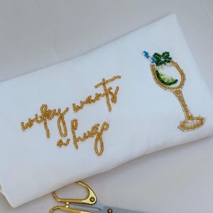 May include: White fabric embroidered with gold beads spelling "Wifey wants a hug" and a cocktail glass design. Gold scissors with silver blades are in the foreground.