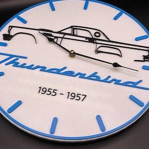 May include: A white and blue wall clock featuring a black outline of a classic car. The clock face includes the word "Thunderbird" in blue script, along with the years "1955 - 1957". The clock has blue hour markers and black clock hands.