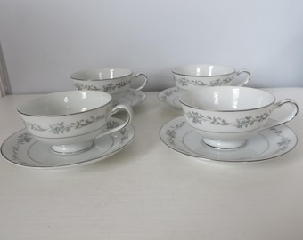 May include: Four white teacups and saucers with a silver trim and a delicate floral pattern. The cups have a classic shape with a handle, and the saucers are round. The set is arranged on a white surface.