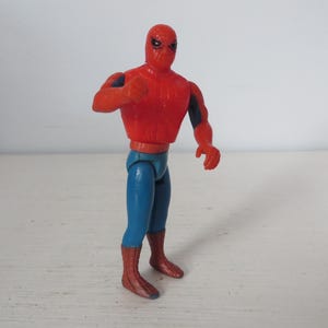 May include: A red and blue plastic action figure of Spider-Man. The figure has a red and blue suit with a spider logo on the chest. The figure is posed with its arms outstretched and its legs slightly bent.