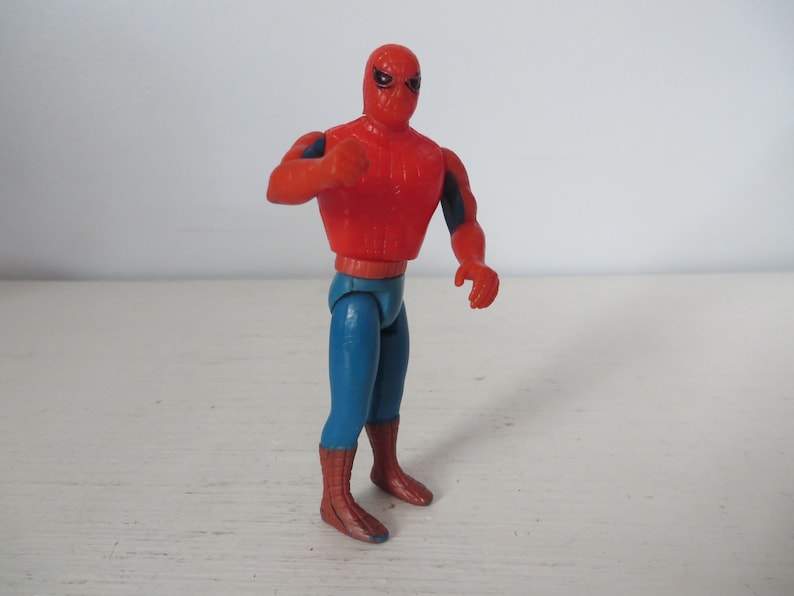 May include: A red and blue plastic action figure of Spider-Man. The figure has a red and blue suit with a spider logo on the chest. The figure is posed with its arms outstretched and its legs slightly bent.