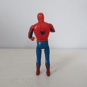 May include: A red and blue plastic action figure of Spider-Man, standing with its back to the camera. The figure has a black spider symbol on its chest.