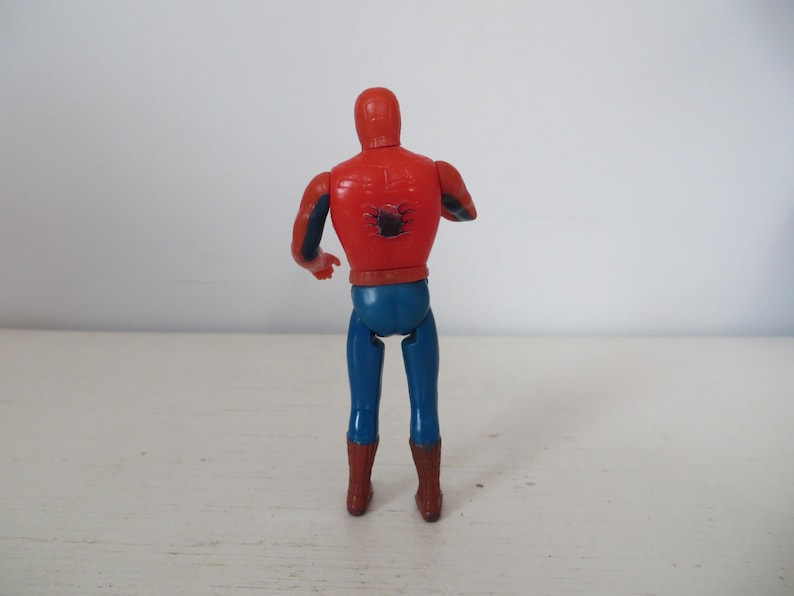 May include: A red and blue plastic action figure of Spider-Man, standing with its back to the camera. The figure has a black spider symbol on its chest.