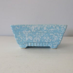 May include: A rectangular, light blue ceramic planter with a speckled white pattern. The planter has a slightly trapezoidal shape, with a ridged design on the lower front and sits on four small feet. A decorative item.