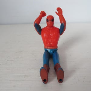 May include: A red and blue plastic action figure of Spider-Man with his arms raised in the air. The figure has a red and blue suit with a spider logo on the chest.