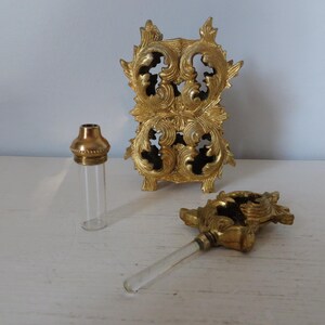 May include: A vintage perfume bottle set with a gold-toned metal stand and applicator. The set includes a clear glass bottle with a gold cap, a decorative stand, and a glass applicator with a gold handle. The stand has an ornate, scrolled design.