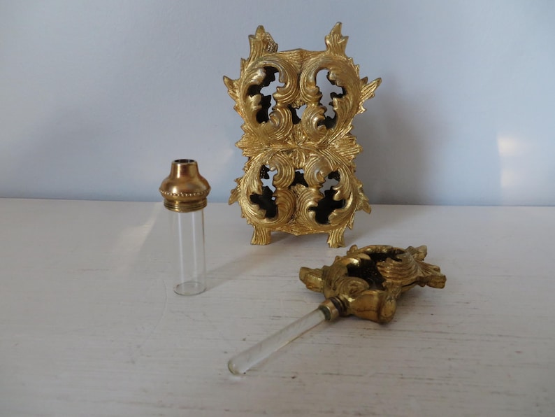 May include: A vintage perfume bottle set with a gold-toned metal stand and applicator. The set includes a clear glass bottle with a gold cap, a decorative stand, and a glass applicator with a gold handle. The stand has an ornate, scrolled design.