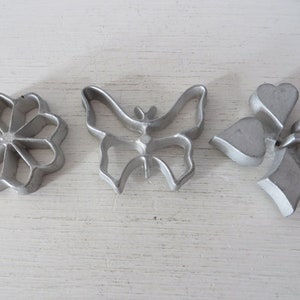May include: Three metal cookie cutters in the shapes of a flower, butterfly, and a set of playing card suits. The cutters are silver in color and have a shiny finish.