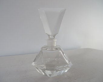 May include: A clear glass bottle with a geometric, faceted design. The bottle has a square base and a tall, pointed stopper.