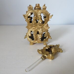 May include: Ornate gold-colored perfume bottle with a matching stopper. The bottle has a decorative, openwork design with floral and scroll motifs. The stopper has a similar design with a clear glass applicator.