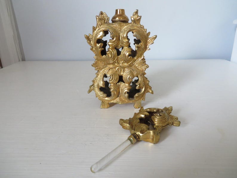 May include: Ornate gold-colored perfume bottle with a matching stopper. The bottle has a decorative, openwork design with floral and scroll motifs. The stopper has a similar design with a clear glass applicator.