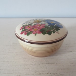 May include: A small, round ceramic trinket box with a lid. The lid is decorated with a floral design featuring pink, blue, and white flowers. The box has a brown rim and a light brown base.