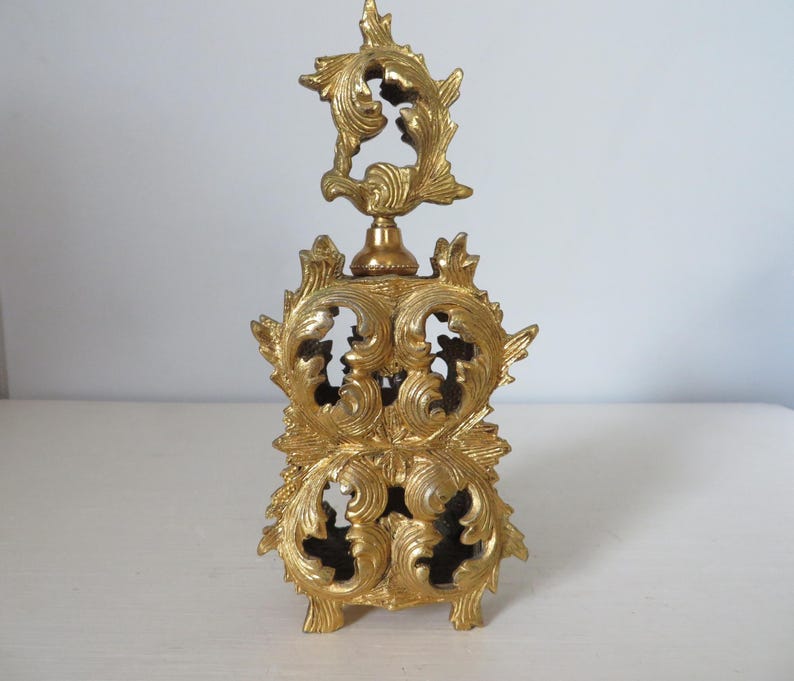 May include: Ornate gold-colored perfume bottle with intricate, swirling leaf and floral designs. The bottle has a decorative top and a rectangular base. The overall design is vintage and elegant, suitable for display or use.