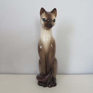 May include: A ceramic Siamese cat figurine in a seated position. The cat has a light brown body, dark brown ears, face, and paws, and blue eyes. The figurine is approximately 12 inches tall and has a glossy finish.