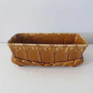 May include: A brown ceramic planter with a rectangular shape and a decorative design of arches and leaves. The planter has four feet and is designed to hold plants.