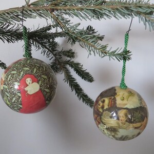 May include: Two round Christmas ornaments hanging from a pine branch. The ornaments have a vintage design with images of people in red and cream-colored clothing. The ornaments are suspended by green twine.