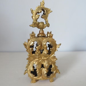 May include: A gold-colored, ornate perfume bottle with a detailed, openwork design. The bottle features a decorative top and base with swirling, leaf-like patterns. The overall aesthetic is vintage and elegant, suitable for display or use.