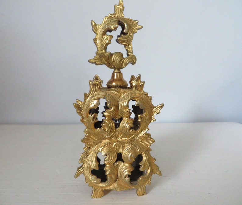 May include: A gold-colored, ornate perfume bottle with a detailed, openwork design. The bottle features a decorative top and base with swirling, leaf-like patterns. The overall aesthetic is vintage and elegant, suitable for display or use.
