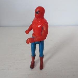 May include: A red and blue plastic action figure of Spider-Man. The figure has a red and blue suit with a spider logo on the chest. The figure is posed in a standing position with its arms raised.