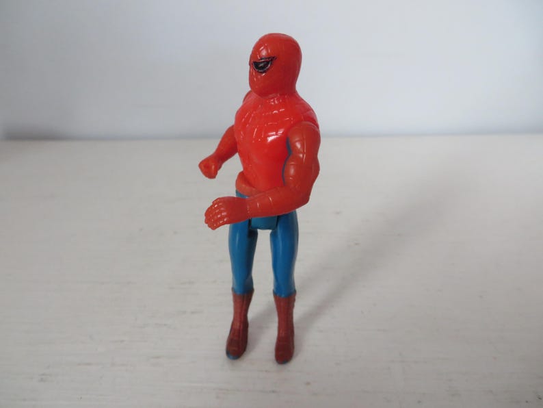 May include: A red and blue plastic action figure of Spider-Man. The figure has a red and blue suit with a spider logo on the chest. The figure is posed in a standing position with its arms raised.