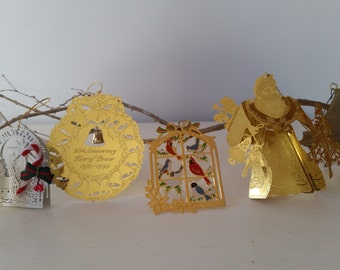 May include: A collection of Christmas ornaments in various shapes and designs, including an angel, a bell, a window with birds, a Santa Claus figure, and a piano. The ornaments are gold-colored and are displayed on a branch.