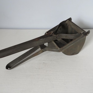 May include: An antique, metal potato ricer with a dark, aged finish. The ricer features a long handle, a basket with a mesh bottom, and a lever mechanism. This kitchen utensil is designed for mashing potatoes and other soft foods.