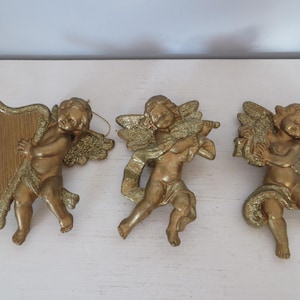 May include: Three gold-colored cherub figurines. One cherub holds a harp, another a violin, and the third a bundle of wheat. Each cherub has detailed wings and is posed in a dynamic stance. The figurines are likely decorative ornaments.