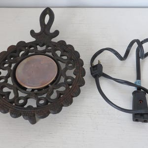 May include: An antique cast iron trivet with an ornate, openwork design and a central, circular ceramic insert. A black electrical cord with a plug is next to the trivet. The trivet is likely used for placing hot cookware on a table.
