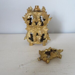 May include: Ornate gold-colored perfume bottle with a matching stopper. The bottle has a detailed, openwork design with floral and scroll motifs. The stopper has a clear glass applicator.