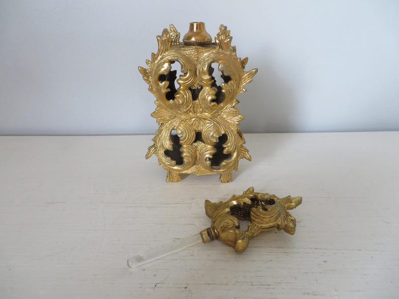 May include: Ornate gold-colored perfume bottle with a matching stopper. The bottle has a detailed, openwork design with floral and scroll motifs. The stopper has a clear glass applicator.