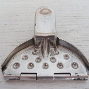 May include: Silver-toned metal citrus juicer with a hinged base and a textured surface. The juicer has a handle on the top and a series of small holes on the bottom.