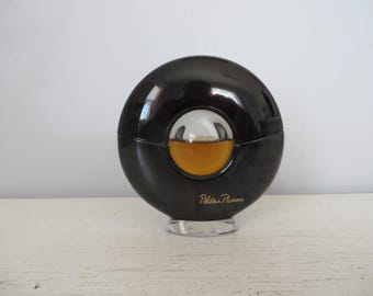 May include: A black and gold round perfume bottle with a clear glass center and the text "Paloma Picasso" on the bottom.