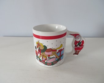 May include: White ceramic Christmas mug with a red stripe and a Santa Claus handle. The mug features illustrations of a rocking horse, a toy soldier, and wrapped gifts. The background has a pattern of small gray dots.