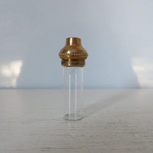 May include: A small, clear glass vial with a decorative gold-colored metal cap. The cap has a rounded top and a beaded design around the base. The vial is empty and sits on a white surface.