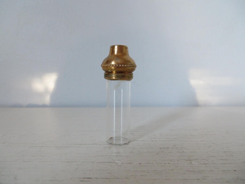 May include: A small, clear glass vial with a decorative gold-colored metal cap. The cap has a rounded top and a beaded design around the base. The vial is empty and sits on a white surface.