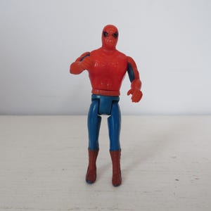 May include: A red and blue plastic action figure of Spider-Man. The figure has a red and blue suit with a spider logo on the chest. The figure is posed with its arms outstretched and its legs slightly bent.