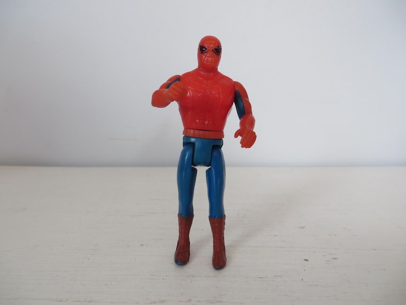 May include: A red and blue plastic action figure of Spider-Man. The figure has a red and blue suit with a spider logo on the chest. The figure is posed with its arms outstretched and its legs slightly bent.