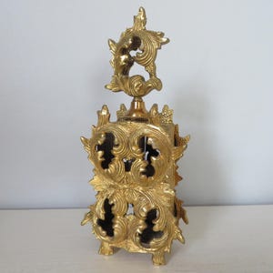 May include: Ornate gold-colored perfume bottle with intricate, swirling leaf and floral designs. The bottle has a rectangular base and a tall, decorative stopper. The design features cut-out sections, adding depth and visual interest to the piece.