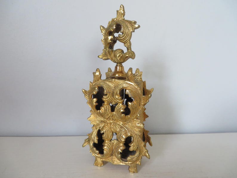 May include: Ornate gold-colored perfume bottle with intricate, swirling leaf and floral designs. The bottle has a rectangular base and a tall, decorative stopper. The design features cut-out sections, adding depth and visual interest to the piece.