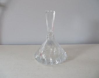 May include: A clear glass perfume bottle with a diamond-shaped base and a tall, slender neck. The bottle has a label that reads "ILLUSIONS 2 oz.  Perfume Oil  Importad by  Avon Products, Inc.  New York, NY".