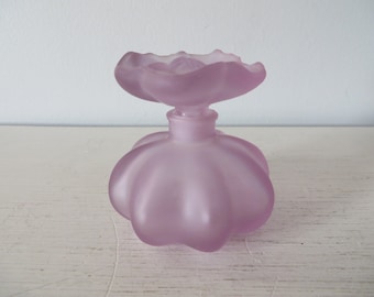 May include: A frosted glass perfume bottle with a flower-shaped stopper. The bottle is a pale lavender color and has a rounded, bulbous shape.