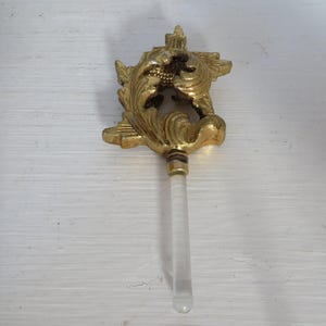 May include: Ornate gold-colored bottle stopper with a decorative floral design. The stopper has a clear, cylindrical handle. A vintage-style decorative item.