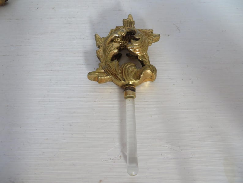 May include: Ornate gold-colored bottle stopper with a decorative floral design. The stopper has a clear, cylindrical handle. A vintage-style decorative item.