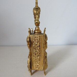 May include: A gold-colored, ornate perfume bottle with a detailed, rectangular base and a decorative top. The bottle features intricate floral and scrollwork patterns, with a bird-like figure atop the stopper. The bottle stands on four small feet.