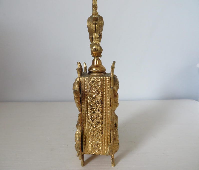 May include: A gold-colored, ornate perfume bottle with a detailed, rectangular base and a decorative top. The bottle features intricate floral and scrollwork patterns, with a bird-like figure atop the stopper. The bottle stands on four small feet.