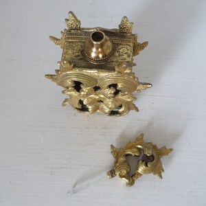 May include: An antique gold-colored perfume bottle with ornate floral and scrollwork detailing. The bottle is square-shaped with a rounded top and a separate decorative stopper with a clear glass applicator.