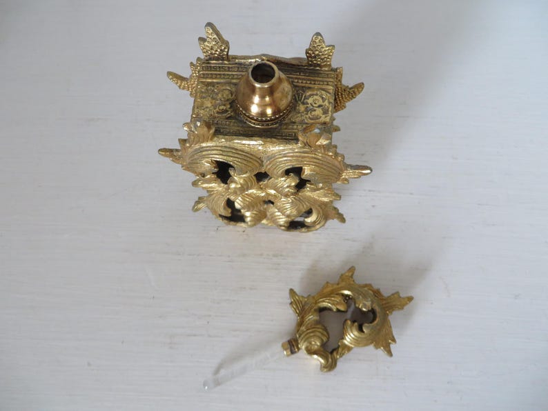 May include: An antique gold-colored perfume bottle with ornate floral and scrollwork detailing. The bottle is square-shaped with a rounded top and a separate decorative stopper with a clear glass applicator.