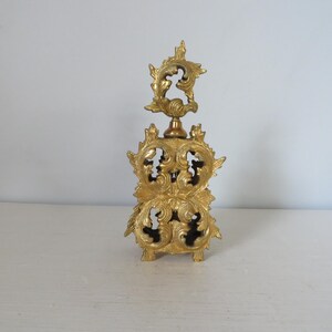 May include: A gold-colored, ornate perfume bottle with a decorative, openwork design. The bottle features a detailed, floral-inspired pattern and a star-shaped stopper. The bottle is made of metal and sits on a white surface.