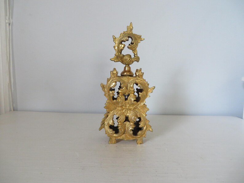May include: A gold-colored, ornate perfume bottle with a decorative, openwork design. The bottle features a detailed, floral-inspired pattern and a star-shaped stopper. The bottle is made of metal and sits on a white surface.